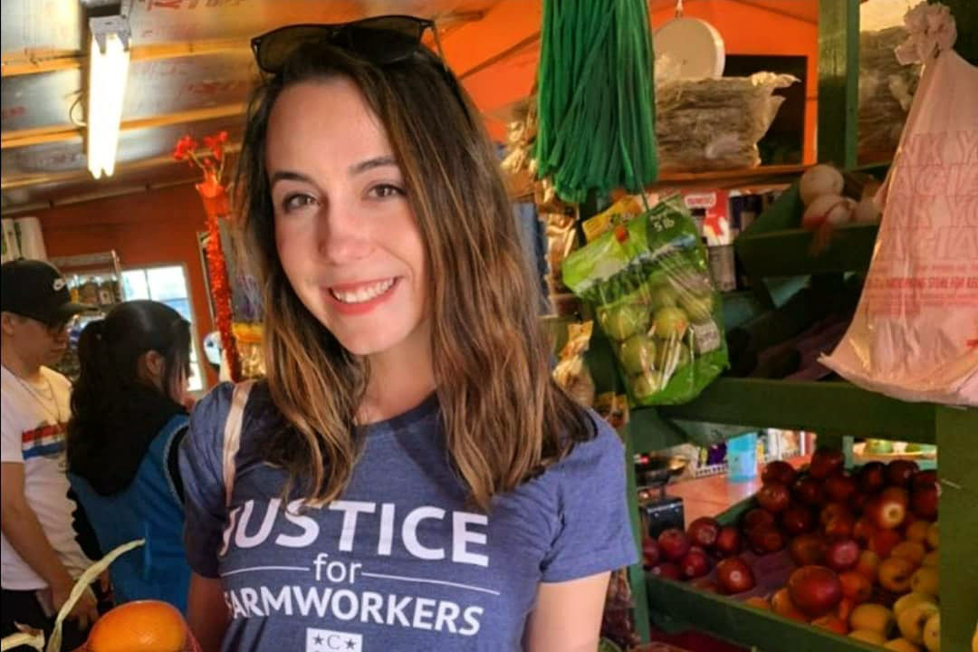 McKenzie Swain standing at a farmers market with a t-shirt reading "Justice for Farm Workers"